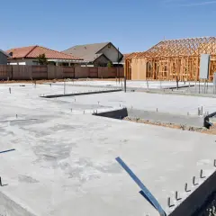 Fundamenty domu w budowie. Widać betonowe płyty i zbrojenie. W tle drewniana konstrukcja domu i inne budynki. Chudy beton proporcje są kluczowe dla trwałości.
