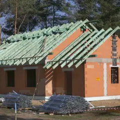 Budowa domu metodą gospodarczą w toku. Dom z cegły, z zieloną więźbą dachową, otoczony materiałami budowlanymi.