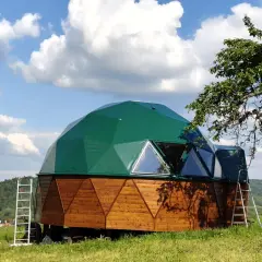 Zielony, nowoczesny dom kopułowy z drewnianą podstawą, otoczony zieloną trawą i drzewami.